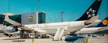 An airplane is parked on an airport tarmac, with its engines and landing gear visible. The airplane has a white body with a logo on its tail fin. An aircraft boarding stair is positioned next to the plane, and there are small vehicles and equipment scattered around, suggesting ground operations. The backdrop includes an airport terminal building with clear skies above.