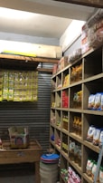 A small, organized storage room lined with wooden shelves filled with various packaged goods, including instant noodles, snacks, and cooking oils. On the left, there are cardboard boxes on a wooden table and a wall lined with hanging packs. The room has a rustic and utilitarian appearance.