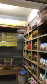 A small, organized storage room lined with wooden shelves filled with various packaged goods, including instant noodles, snacks, and cooking oils. On the left, there are cardboard boxes on a wooden table and a wall lined with hanging packs. The room has a rustic and utilitarian appearance.