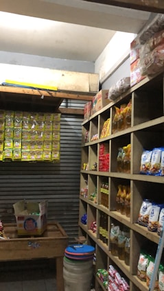 A small, organized storage room lined with wooden shelves filled with various packaged goods, including instant noodles, snacks, and cooking oils. On the left, there are cardboard boxes on a wooden table and a wall lined with hanging packs. The room has a rustic and utilitarian appearance.