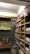 A small, organized storage room lined with wooden shelves filled with various packaged goods, including instant noodles, snacks, and cooking oils. On the left, there are cardboard boxes on a wooden table and a wall lined with hanging packs. The room has a rustic and utilitarian appearance.