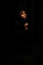 A woman is depicted with a soft expression, eyes closed, hands clasped together. Her head is covered with a dark shawl, and she is set against a black background, creating a serene and contemplative atmosphere.