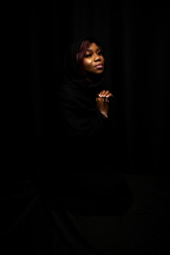 A woman is depicted with a soft expression, eyes closed, hands clasped together. Her head is covered with a dark shawl, and she is set against a black background, creating a serene and contemplative atmosphere.