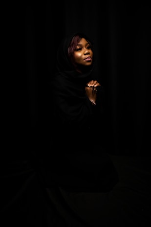 A woman is depicted with a soft expression, eyes closed, hands clasped together. Her head is covered with a dark shawl, and she is set against a black background, creating a serene and contemplative atmosphere.