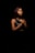 A woman stands against a dark background, with her eyes closed and hands clasped together in a gesture of prayer or contemplation. She wears a black, sleeveless top with a sequined pattern on it.