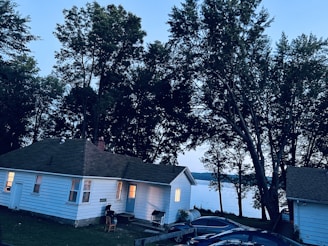 A cozy cottage exterior at dusk with warm lights glowing through the windows, surrounded by lush greenery.