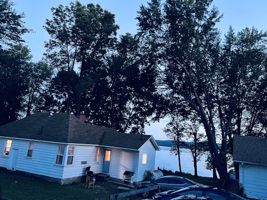 A small white cottage with illuminated windows is nestled among tall trees during twilight. The house is surrounded by a lush green lawn and several parked cars. The backdrop features a calm lake partially visible through the trees, creating a serene and peaceful atmosphere.
