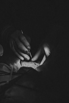 A black and white photograph captures a close-up of a person's hands wearing gloves, holding a surgical instrument over a surface, possibly during a medical procedure.