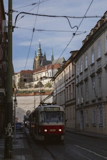 A streetcar travels down a narrow, cobblestone street lined with historic buildings. Overhead tram wires crisscross the scene. In the background, a grand castle with tall spires rises against a clear sky, adding a sense of historical ambiance.