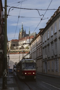 A streetcar travels down a narrow, cobblestone street lined with historic buildings. Overhead tram wires crisscross the scene. In the background, a grand castle with tall spires rises against a clear sky, adding a sense of historical ambiance.