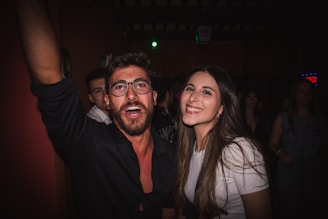 A group of young adults enjoying themselves at a social event or party indoors. The man on the left is wearing glasses and a dark shirt, looking cheerful, while the woman on the right, with long brown hair, smiles brightly. Several other people are visible in the dimly lit background.