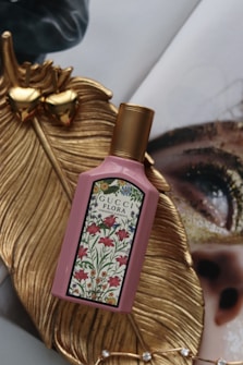 A bottle of Gucci Flora Gorgeous Gardenia perfume is placed on a decorative golden leaf. The bottle is pink with a detailed floral pattern on the front label. In the background, there is a close-up of a fashion magazine featuring an eye with gold glitter makeup, creating a luxurious setting.