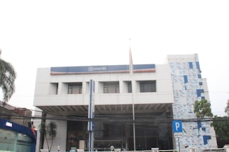 A modern, multi-story building with a predominantly white exterior and a blue sign prominently displaying 'Bank BRI'. The facade has rectangular windows and a small flagpole in front. There is vegetation including palm trees to the side and a parking sign visible on the street.