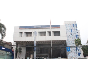 A modern, multi-story building with a predominantly white exterior and a blue sign prominently displaying 'Bank BRI'. The facade has rectangular windows and a small flagpole in front. There is vegetation including palm trees to the side and a parking sign visible on the street.