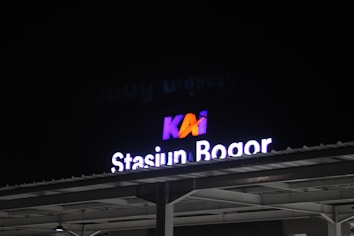 A sign for 'Stasiun Bogor' is illuminated against a dark night sky. The letters are bright and stand out, with the 'KAI' logo in contrasting colors above them. The structure of the station extends below the sign.