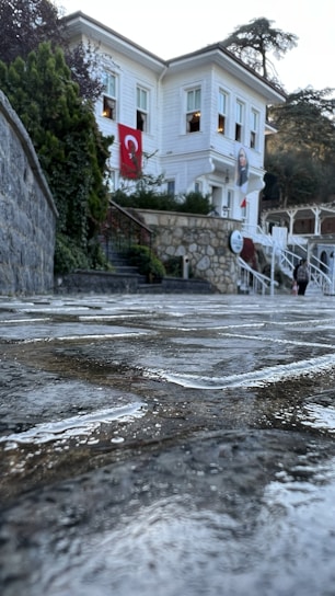 A white two-story building with a Turkish flag hanging from a window. The building is surrounded by greenery and stone walls. The foreground features a wet, reflective stone pathway, suggesting recent rain.