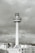 A tall, cylindrical tower rises above a cityscape, topped with a large broadcasting structure. The structure has a bold, circular band displaying radio station details. The tower is surrounded by a group of lower buildings with a cloudy sky in the background.