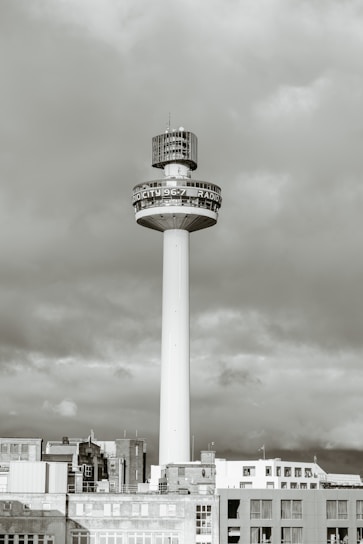 A tall, cylindrical tower rises above a cityscape, topped with a large broadcasting structure. The structure has a bold, circular band displaying radio station details. The tower is surrounded by a group of lower buildings with a cloudy sky in the background.