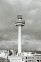 A tall, cylindrical tower rises above a cityscape, topped with a large broadcasting structure. The structure has a bold, circular band displaying radio station details. The tower is surrounded by a group of lower buildings with a cloudy sky in the background.