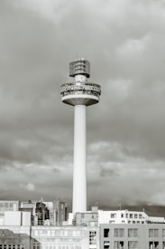 A tall, cylindrical tower rises above a cityscape, topped with a large broadcasting structure. The structure has a bold, circular band displaying radio station details. The tower is surrounded by a group of lower buildings with a cloudy sky in the background.