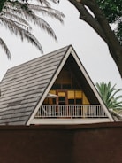 A triangular A-frame house with a steep roof and a balcony featuring a chair and umbrella. The structure is framed by palm tree leaves, suggesting a tropical setting.