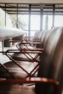 A row of modern chairs with metal armrests aligned along a sleek conference table inside a room with large windows. The room is well-lit with natural light streaming in from outside, where greenery can be seen.