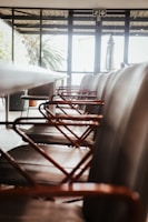 A row of modern chairs with metal armrests aligned along a sleek conference table inside a room with large windows. The room is well-lit with natural light streaming in from outside, where greenery can be seen.