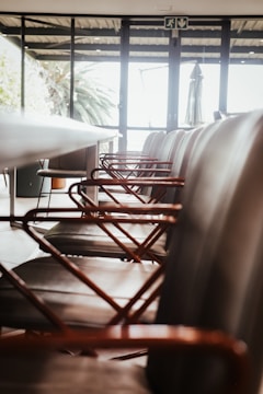 A row of modern chairs with metal armrests aligned along a sleek conference table inside a room with large windows. The room is well-lit with natural light streaming in from outside, where greenery can be seen.