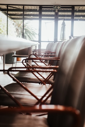 A row of modern chairs with metal armrests aligned along a sleek conference table inside a room with large windows. The room is well-lit with natural light streaming in from outside, where greenery can be seen.