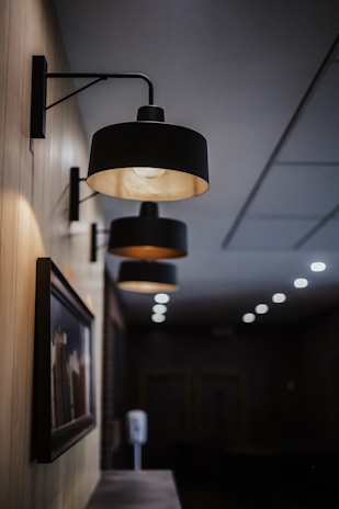 Several wall-mounted black lamps with a modern design are arranged in a linear perspective along a wooden-paneled wall. The lamps are illuminated, casting a warm glow. Below the lamps is a framed picture, slightly blurred, which adds an artistic element to the setting. The background is a dimly lit hallway or lobby area, with a series of small ceiling lights visible further in the distance.