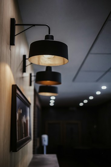 Several wall-mounted black lamps with a modern design are arranged in a linear perspective along a wooden-paneled wall. The lamps are illuminated, casting a warm glow. Below the lamps is a framed picture, slightly blurred, which adds an artistic element to the setting. The background is a dimly lit hallway or lobby area, with a series of small ceiling lights visible further in the distance.