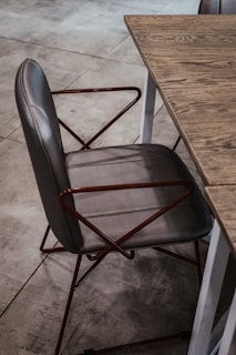 A stylish gray chair with a leather seat and backrest, featuring a geometric metal frame with angled armrests, positioned next to a wooden table with a visible grain pattern on the tabletop. The setting is on a tiled floor, contributing to a modern aesthetic.