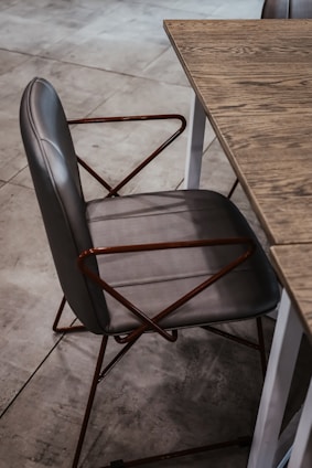 A stylish gray chair with a leather seat and backrest, featuring a geometric metal frame with angled armrests, positioned next to a wooden table with a visible grain pattern on the tabletop. The setting is on a tiled floor, contributing to a modern aesthetic.