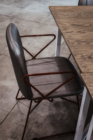 A stylish gray chair with a leather seat and backrest, featuring a geometric metal frame with angled armrests, positioned next to a wooden table with a visible grain pattern on the tabletop. The setting is on a tiled floor, contributing to a modern aesthetic.