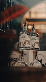 Two bottles of alcoholic beverage stand atop a cardboard box with additional bottles partially visible. The bottles feature labels with text and a graphic design. The setting appears to be a dimly lit area with a warm, muted ambiance, possibly a bar or a store.