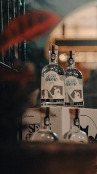 Two bottles of alcoholic beverage stand atop a cardboard box with additional bottles partially visible. The bottles feature labels with text and a graphic design. The setting appears to be a dimly lit area with a warm, muted ambiance, possibly a bar or a store.