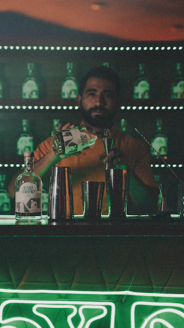 A Latin American bartender mid-pour, sending a cascade of amber cocktail into a glass, backlit by moody, deep tones