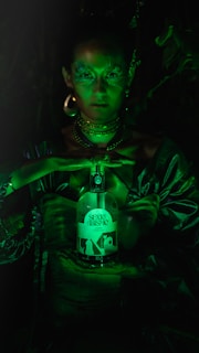 A person with shimmering green makeup and elaborate jewelry holds a bottle labeled 'Sexto Abrismo'. The scene is dimly lit with a green hue, giving it a mystical and otherworldly appearance.