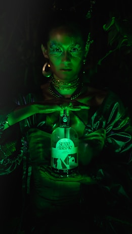 A person with shimmering green makeup and elaborate jewelry holds a bottle labeled 'Sexto Abrismo'. The scene is dimly lit with a green hue, giving it a mystical and otherworldly appearance.