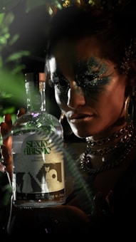 A person with intricate facial makeup and elaborate jewelry is holding a bottle labeled 'Sexto Abismo'. The scene has a mysterious, dimly lit atmosphere with green foliage partially obscuring the view.