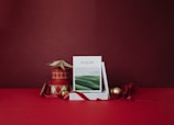 A stack of books with a cover titled 'PSALMS' featuring a serene landscape, is positioned on a red surface. Next to it, there are decorative items such as circular boxes with red and cream diamond patterns, a star-shaped ornament, golden and red Christmas baubles, and a ribbon. The background is a rich red color, creating an overall festive atmosphere.