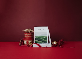 A stack of books with a cover titled 'PSALMS' featuring a serene landscape, is positioned on a red surface. Next to it, there are decorative items such as circular boxes with red and cream diamond patterns, a star-shaped ornament, golden and red Christmas baubles, and a ribbon. The background is a rich red color, creating an overall festive atmosphere.