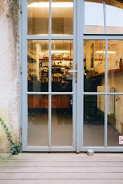 A glass door with blue-gray frames and multiple panes leads into a cozy interior displaying shelves stocked with various items and a counter. The reflection on the glass indicates a wooden deck leading to the door, with a stone propping it slightly open. Part of the exterior wall shows a climbing plant on the left side.