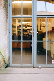 A glass door with blue-gray frames and multiple panes leads into a cozy interior displaying shelves stocked with various items and a counter. The reflection on the glass indicates a wooden deck leading to the door, with a stone propping it slightly open. Part of the exterior wall shows a climbing plant on the left side.