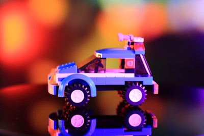 A small, colorful toy car built from Lego bricks is placed against a soft-focus, multicolored bokeh background. The car is primarily blue with black wheels and has various details in white, pink, and transparent bricks. It is reflected on a glossy surface, adding an element of symmetry to the composition.