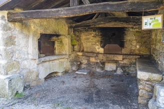 An old stone oven structure is seen inside a rustic building with weathered wooden beams and stone walls. There are two oven openings with darkened interiors, suggesting they have been heavily used. A small informational board is hanging on one side, and the ground is covered with gravel.