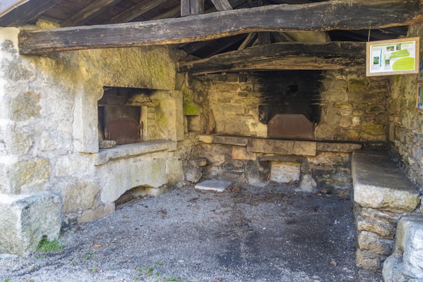 An old stone oven structure is seen inside a rustic building with weathered wooden beams and stone walls. There are two oven openings with darkened interiors, suggesting they have been heavily used. A small informational board is hanging on one side, and the ground is covered with gravel.