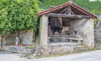 A small rustic stone structure with a wooden roof houses an old-fashioned brick oven. The oven has a large black metal chimney hood. The area around the structure is adorned with lush greenery, including climbing vines. There is a stone bench to the right of the oven and two rectangular planters with red flowers on a concrete platform to the left. A narrow pathway runs in front of the structure.