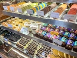 A display case featuring a variety of colorful macarons, pastries, and chocolate-covered treats. The macarons are arranged neatly in rows with labels indicating flavors and prices. Below, there are sections of chocolate eclairs, tarts, and other assorted desserts, showcasing a vibrant and inviting presentation of sweet delicacies.