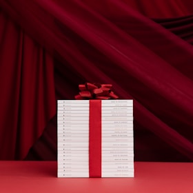A neatly stacked collection of white books is tied together with a bold red ribbon and bow, set against a rich red draped fabric background. The titles on the book spines indicate they are religious texts, adding a reverent and celebratory ambiance.